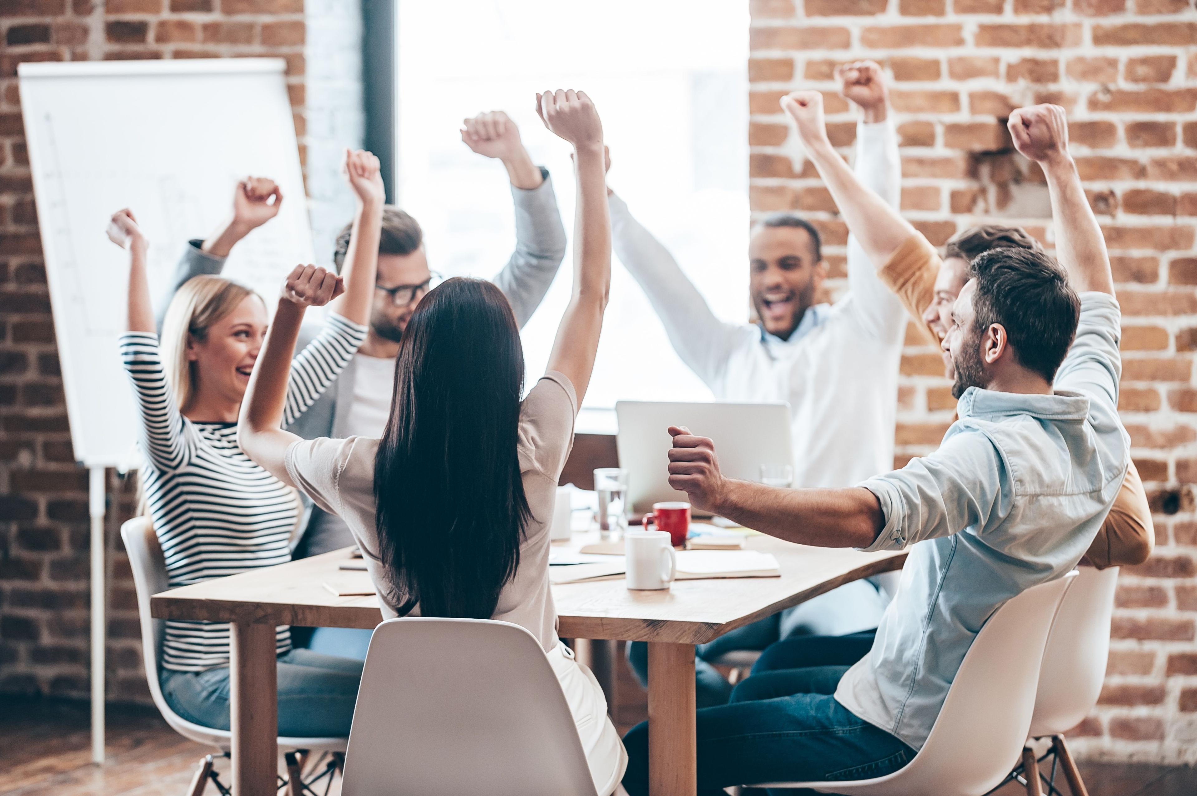 Group of coworkers raising their arms and celebrating together during an office meeting.